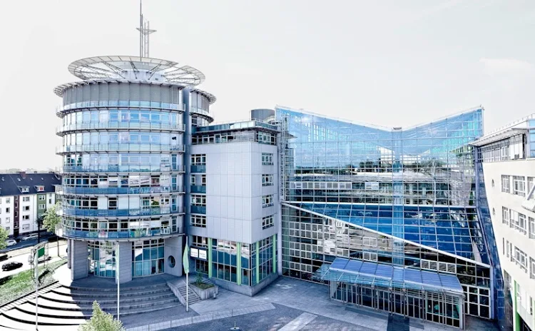 Moderner Gebäudekomplex mit gläsernen Fassaden, rundem Turm und geschwungenen Dächern, umgeben von gepflasterten Plätzen und angrenzenden Wohngebäuden.